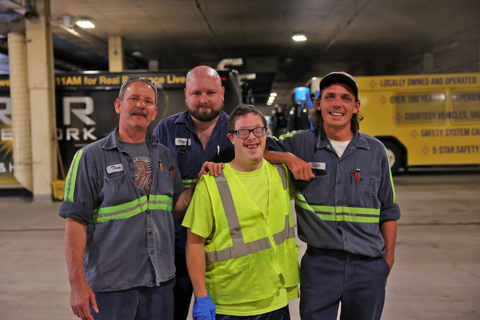 Four coworkers in work uniforms and safety vests stand together smiling in a bus garage